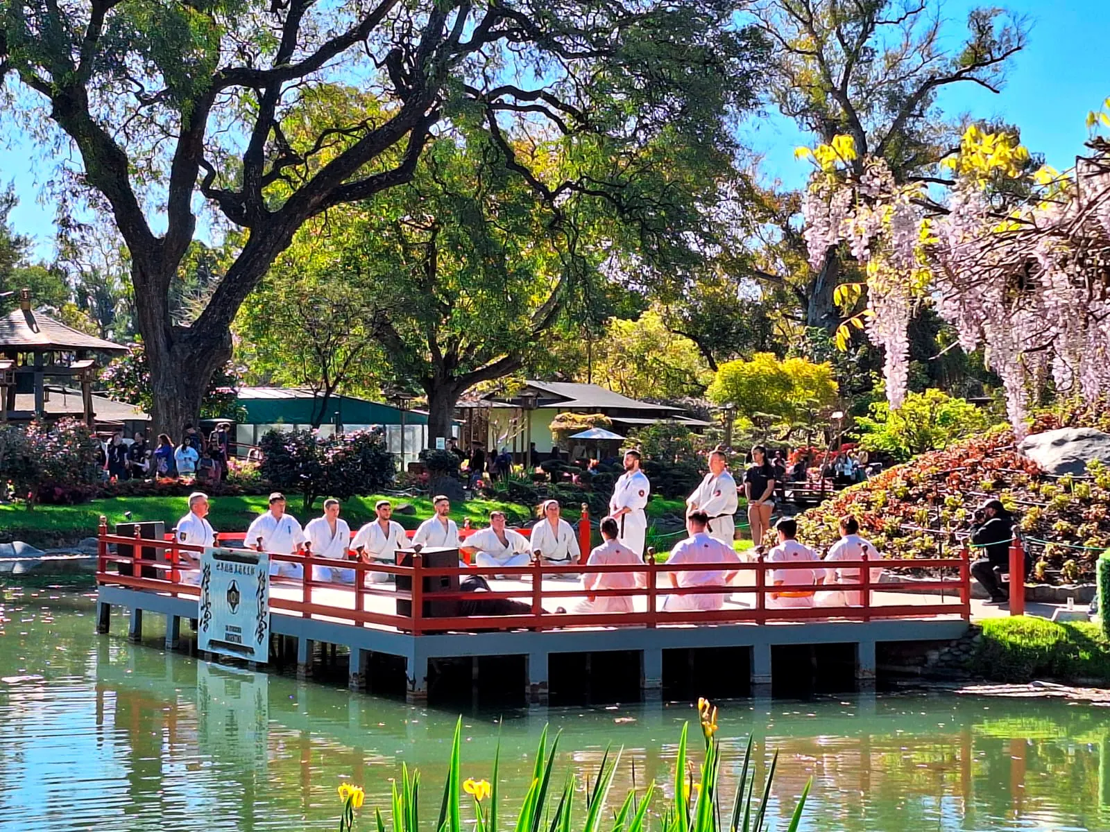 Enbu (demostración) de karate Kyokushin sobre una plataforma junto al lago del Jardín Japonés, con integrantes del dojo sentados en seiza.