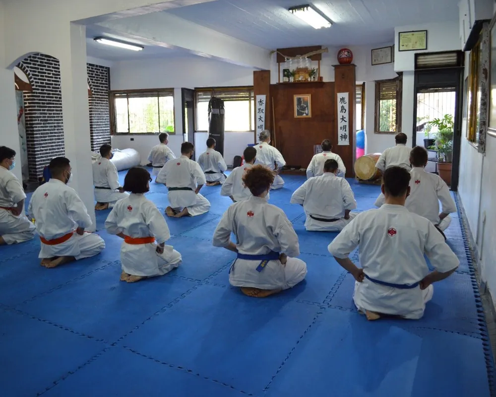Interior del dojo: alumnos de espaldas, sentados en seiza sobre el tatami azul, frente al altar de la pared del fondo.