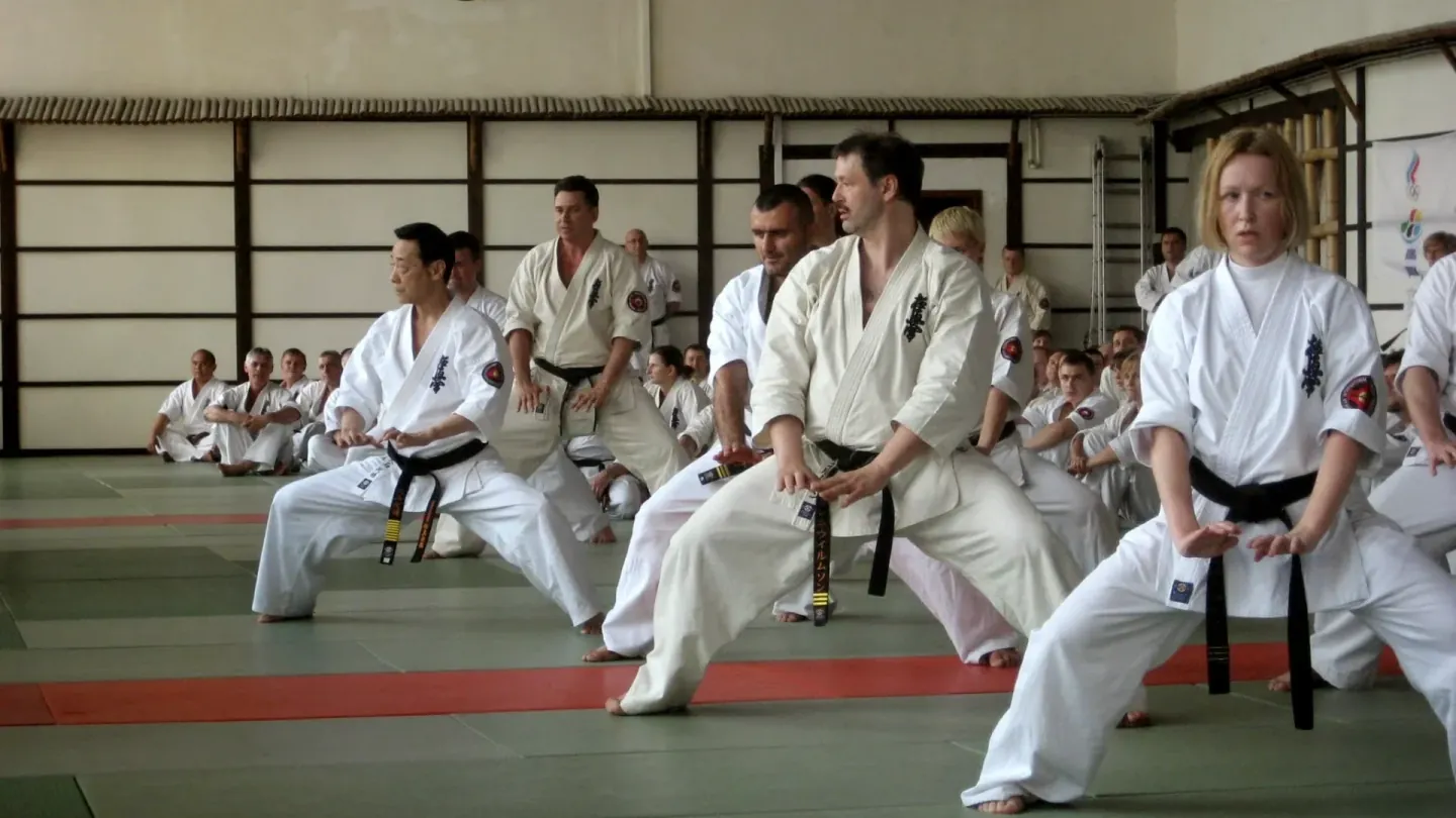 Practicantes de Kyokushin en un dojo, en una misma postura de kata, con otros alumnos sentados detrás.